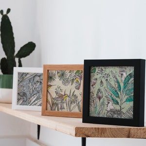 Pode incluir: Três impressões de arte emolduradas com ilustrações botânicas. As molduras são brancas, de madeira natural e pretas. As impressões apresentam desenhos detalhados de plantas, pássaros e animais em uma variedade de cores, incluindo verde, azul e amarelo. As molduras são exibidas em uma prateleira de madeira.