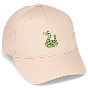 Könnte beinhalten: Eine hellbeige Baseballkappe mit einer Stickerei eines kleinen grünen Schlangenmotivs mit dunkelgrünen Streifen, großen weißen Augen und einer roten Zunge. Die Kappe verfügt über eine strukturierte Krone, einen gebogenen Schirm und gestickte Ösen.