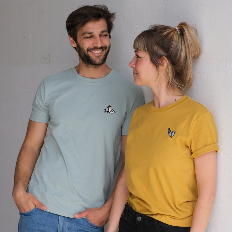 May include: A couple wearing matching t-shirts with embroidered bird and chicken designs. The man is wearing a light blue t-shirt with a white bird holding a cup. The woman is wearing a yellow t-shirt with a gray chicken.