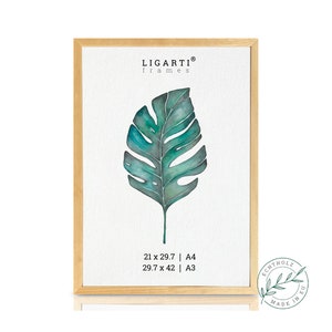Könnte beinhalten: Ein gerahmtes Aquarellbild eines großen grünen Blattes mit einem hellbraunen Holzrahmen. Der Rahmen trägt den Text "LIGARTI® frames" und "21 x 29.7 | A4 29.7 x 42 | A3" auf der Unterseite. Ein runder Stempel mit dem Text "ECHTHOLZ MADE IN EU" befindet sich in der unteren rechten Ecke.
