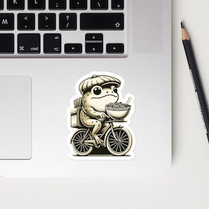 Puede incluir: Una ilustración de dibujos animados de una rana con un sombrero y montando una bicicleta mientras sostiene un tazón de fideos. La rana lleva una bolsa de entrega en la espalda.