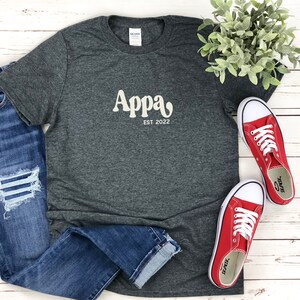 May include: A dark heather gray short-sleeve t-shirt featuring the cream-colored text Appa EST 2022 printed on the chest. The shirt is paired with blue denim jeans that have distressed knee detailing and a pair of bright red low-top sneakers.