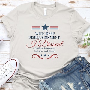 Può includere: T-shirt color crema con la scritta "WITH DEEP DISILLUSIONMENT, I DISSENT" in blu navy e bordeaux. Il design include una stella e strisce. La maglietta è abbinata a pantaloncini di jeans e scarpe da ginnastica bianche.