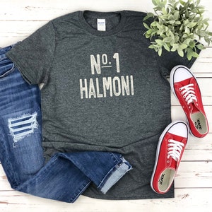 May include: A dark gray t-shirt with the text "No. 1 Halmoni" in a distressed white font. The shirt is paired with blue jeans and red sneakers with white laces. A sprig of greenery is also in the image.