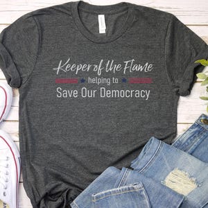May include: A dark grey t-shirt with the text "Keeper of the Flame helping to Save Our Democracy" in white lettering. The text is surrounded by a red, white and blue design that resembles the American flag.