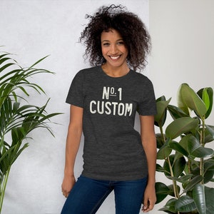 K&ouml;nnte beinhalten: Dunkelgraues T-Shirt mit dem Text "No. 1 Custom" in verblichener wei&szlig;er Schrift. Das Shirt hat einen Rundhalsausschnitt und kurze &Auml;rmel. Das Model tr&auml;gt eine blaue Jeans.
