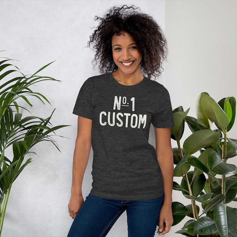K&ouml;nnte beinhalten: Dunkelgraues T-Shirt mit dem Text "No. 1 Custom" in verblichener wei&szlig;er Schrift. Das Shirt hat einen Rundhalsausschnitt und kurze &Auml;rmel. Das Model tr&auml;gt eine blaue Jeans.