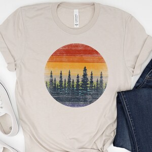 Puede incluir: Una camiseta beige con un gr&aacute;fico de estilo vintage de una puesta de sol detr&aacute;s de una fila de &aacute;rboles. El gr&aacute;fico est&aacute; en un c&iacute;rculo y tiene un aspecto descolorido y desgastado.