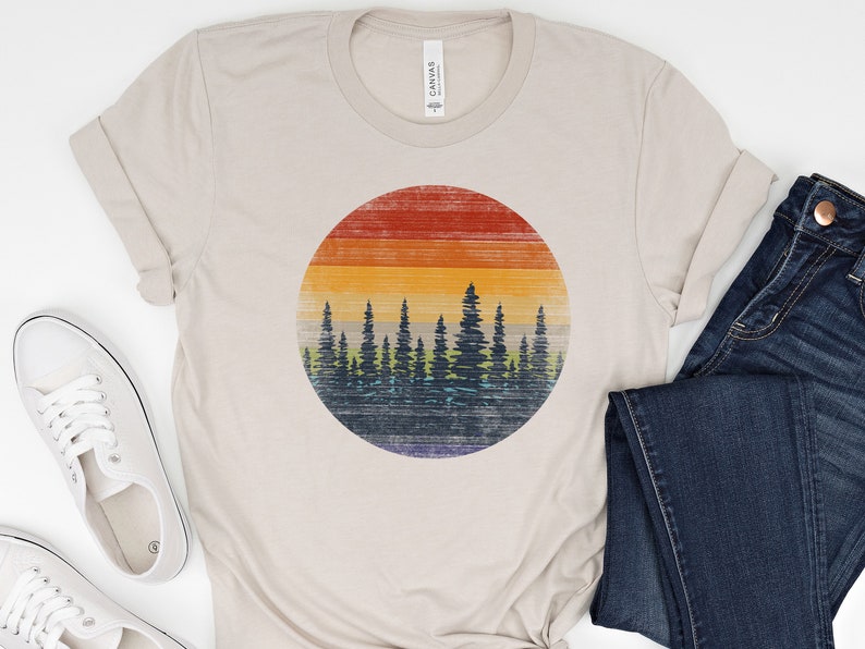 Puede incluir: Una camiseta beige con un gr&aacute;fico de estilo vintage de una puesta de sol detr&aacute;s de una fila de &aacute;rboles. El gr&aacute;fico est&aacute; en un c&iacute;rculo y tiene un aspecto descolorido y desgastado.