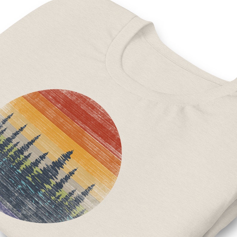 Puede incluir: Una camiseta blanca con un dise&ntilde;o circular que presenta una puesta de sol retro con una silueta de bosque delante. La puesta de sol est&aacute; compuesta por rayas naranja, amarilla, roja y azul.