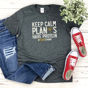 May include: A dark grey t-shirt with the text "Keep Calm Plants Have Protein #PlantStrong" in white and yellow lettering. The shirt is laid out on a white wooden surface with a pair of red trainers and blue jeans.