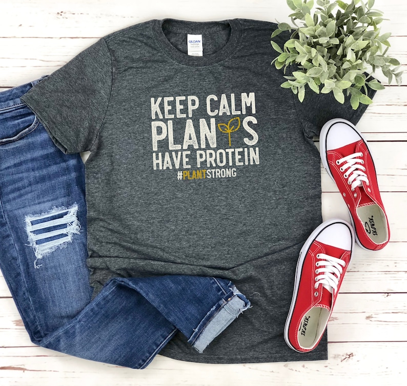 May include: A dark grey t-shirt with the text "Keep Calm Plants Have Protein #PlantStrong" in white and yellow lettering. The shirt is laid out on a white wooden surface with a pair of red trainers and blue jeans.