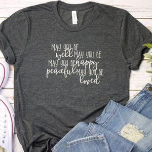 May include: A dark grey t-shirt with the text "May you be well, may you be happy, peaceful, may you be loved" printed in white.