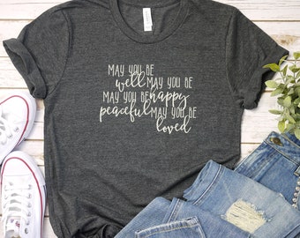 Può includere: Una maglietta grigio scuro con il testo "May you be well, may you be happy, peaceful, may you be loved" stampato in bianco.
