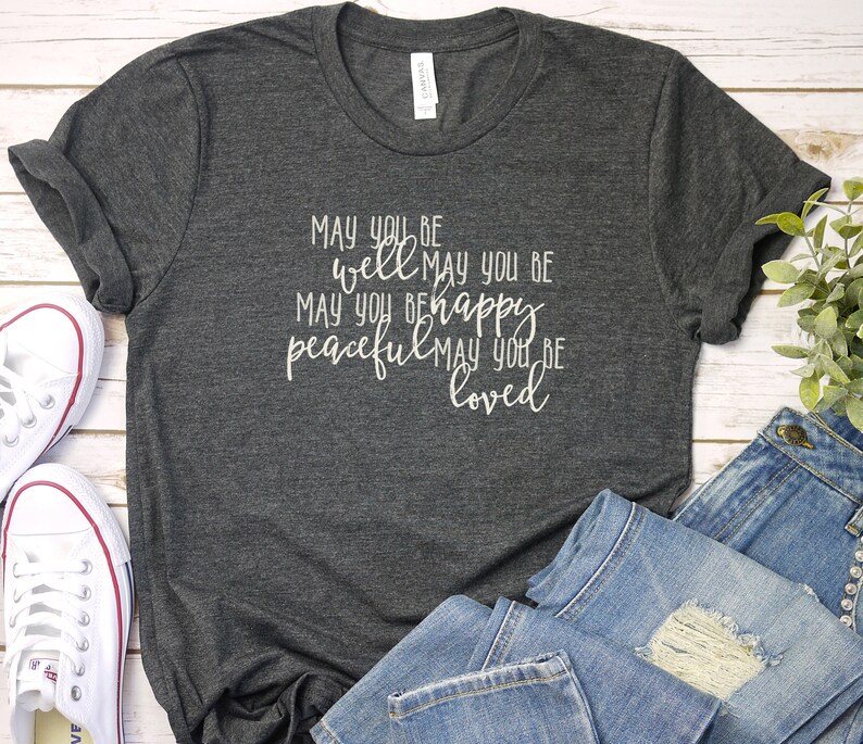 May include: A dark grey t-shirt with the text "May you be well, may you be happy, peaceful, may you be loved" printed in white.