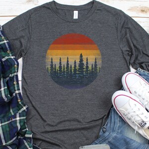 Pu&ograve; includere: Maglietta a maniche lunghe grigio scuro con una grafica circolare di un tramonto su una foresta. Il design ha uno stile retr&ograve; con tonalit&agrave; arancioni, gialle e blu. Sono visibili anche scarpe da ginnastica bianche e una camicia a quadri.