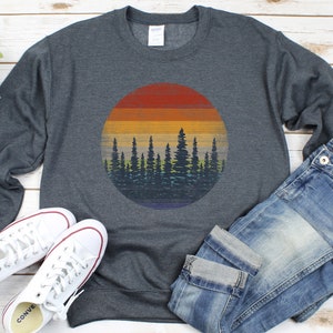 Pu&ograve; includere: Felpa grigio scuro con grafica circolare di un tramonto su una foresta. Il tramonto presenta strisce orizzontali arancioni, gialle e blu. Sono presenti anche scarpe da ginnastica bianche e jeans blu.