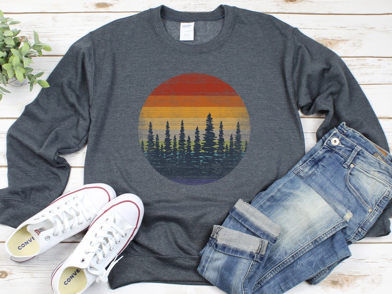 Pu&ograve; includere: Felpa grigio scuro con grafica circolare di un tramonto su una foresta. Il tramonto presenta strisce orizzontali arancioni, gialle e blu. Sono presenti anche scarpe da ginnastica bianche e jeans blu.