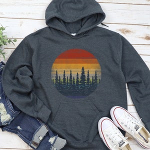 Pu&ograve; includere: Una felpa con cappuccio grigio scuro con una grafica circolare di un tramonto su una foresta. Il design del tramonto presenta bande di rosso, arancione, giallo e blu. La silhouette della foresta &egrave; blu scuro. Scarpe da ginnastica bianche e jeans blu sono accanto.