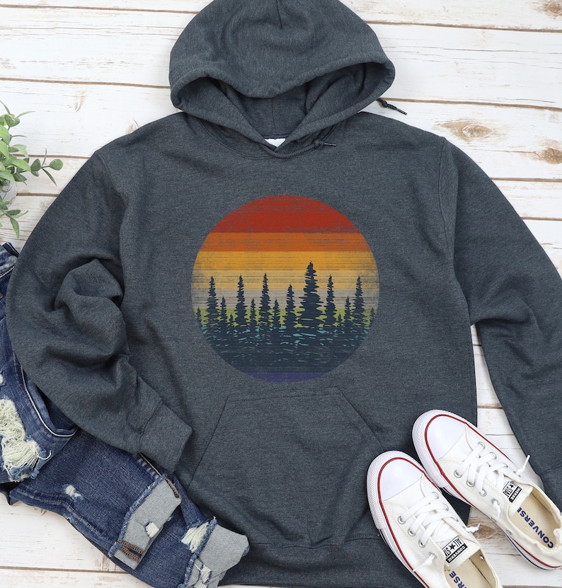 Pu&ograve; includere: Una felpa con cappuccio grigio scuro con una grafica circolare di un tramonto su una foresta. Il design del tramonto presenta bande di rosso, arancione, giallo e blu. La silhouette della foresta &egrave; blu scuro. Scarpe da ginnastica bianche e jeans blu sono accanto.