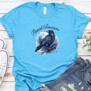 Puede incluir: Camiseta azul claro con un diseño de un cuervo negro posado en una rama, rodeado de un diseño de cielo estrellado. Las palabras "Resist Fascism" están escritas encima del cuervo en cursiva. La camiseta se muestra con pantalones cortos vaqueros.