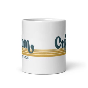 May include: White ceramic mug with a gold and blue design. The mug has the text "Custom" and "Est 2022" on it.