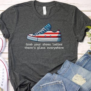 May include: A dark gray t-shirt with a graphic of a red, white, and blue American flag-themed sneaker. The text "Grab your shoes ladies there's glass everywhere" is printed below the sneaker.