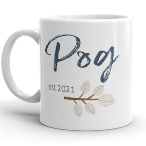 Può includere: Tazza in ceramica bianca con la parola "Pog" scritta a mano in blu e il testo "est 2021" sotto. Un ramo marrone con foglie beige si trova sul lato destro della tazza.