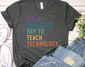 May include: Dark grey t-shirt with the text "IT'S A BEAUTIFUL DAY TO TEACH TECHNOLOGY" in purple, teal, yellow, and orange. The shirt is paired with blue jeans and white canvas trainers.