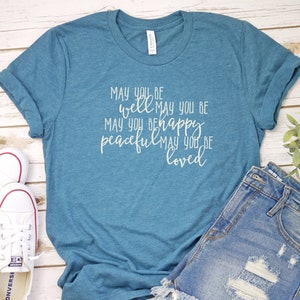 May include: A teal blue t-shirt with the text "MAY YOU BE well MAY YOU BE MAY YOU BE happy peaceful MAY YOU BE loved" printed in white on the front.