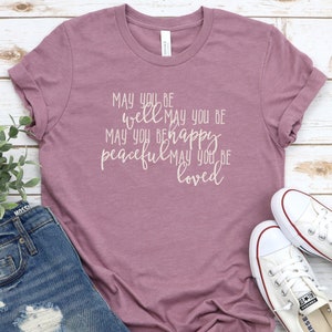 May include: A mauve t-shirt with the text "May you be well May you be happy peaceful May you be loved" printed in white.
