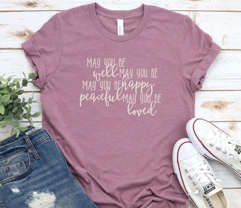 May include: A mauve t-shirt with the text "May you be well May you be happy peaceful May you be loved" printed in white.