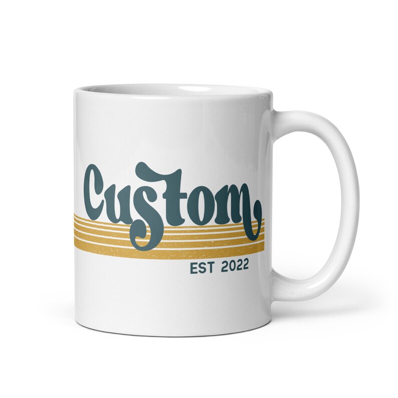 May include: White ceramic mug with a teal blue and gold design. The mug has the word "Custom" in a retro font with gold stripes and the text "EST 2022" below.