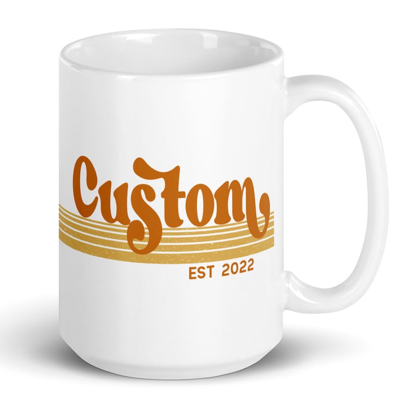 May include: White ceramic coffee mug with a brown and gold design. The mug features the word "Custom" in a retro font with gold stripes and the text "EST 2022" below.