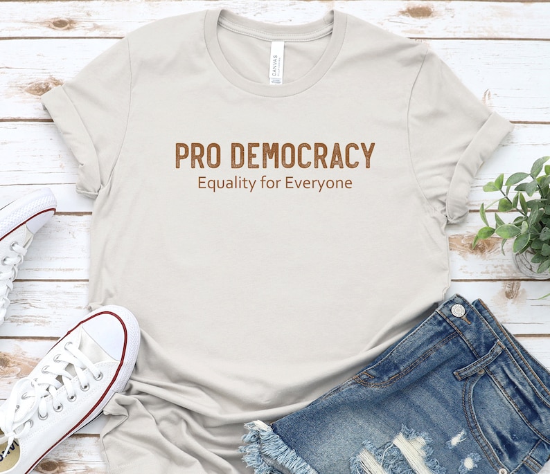May include: A light gray t-shirt with the text "PRO DEMOCRACY" and "Equality for Everyone" in brown lettering.