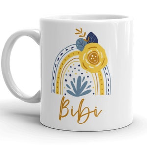 May include: White ceramic mug with a rainbow design in yellow, blue, and white. The rainbow has a yellow flower with brown accents and blue leaves on top. The word "Bibi" is written in gold script below the rainbow.