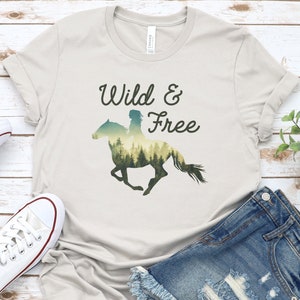 Könnte beinhalten: Ein hellbeiges T-Shirt mit einem Aufdruck eines Pferdes und Reiters, die vor einem Waldmotiv silhouettiert sind. Der Text "Wild & Free" ist in Schwarz über dem Motiv gedruckt.