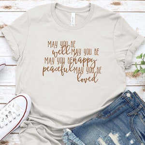 May include: A light grey t-shirt with a brown graphic that reads "MAY YOU BE well MAY YOU BE happy peaceful MAY YOU BE loved".