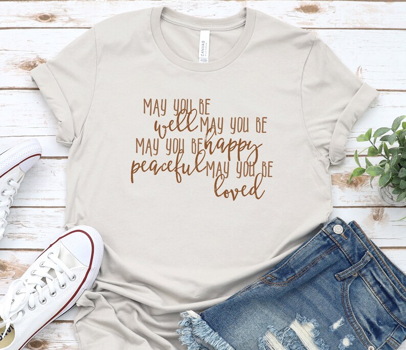 May include: A light grey t-shirt with a brown graphic that reads "MAY YOU BE well MAY YOU BE happy peaceful MAY YOU BE loved".