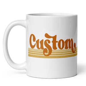 May include: White ceramic coffee mug with the word "Custom" in orange lettering with a retro style font. The word is centered on the mug and is above four thin, horizontal, gold stripes.