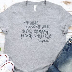 May include: Grey t-shirt with the text "MAY YOU BE well MAY YOU BE MAY YOU BE happy peaceful MAY YOU BE loved" printed on it.