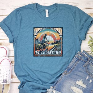 May include: A teal blue t-shirt with a vintage-style graphic of a woman riding a bicycle with the text "Adventure Awaits".