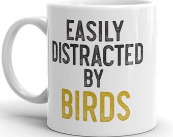 Puede incluir: Taza de cerámica blanca con texto negro que dice "Easily Distracted by Birds" en una fuente desgastada. La palabra "Birds" está en dorado.