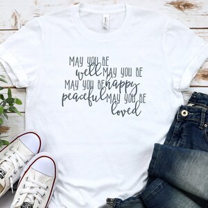 May include: White t-shirt with a grey graphic that reads "May you be well May you be happy peaceful May you be loved".