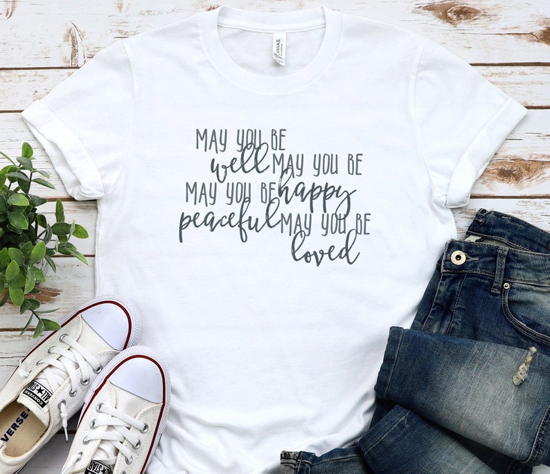 May include: White t-shirt with a grey graphic that reads "May you be well May you be happy peaceful May you be loved".