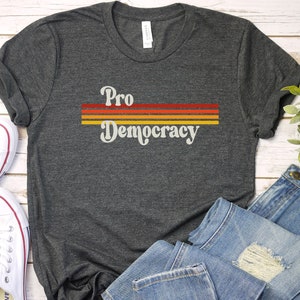 K&ouml;nnte beinhalten: Dunkelgraues T-Shirt mit dem Schriftzug "Pro Democracy" in Retro-Schrift, &uuml;ber drei horizontalen Streifen in Rot, Orange und Gelb. Das Shirt wird mit wei&szlig;en Turnschuhen und blauen Jeans gezeigt.