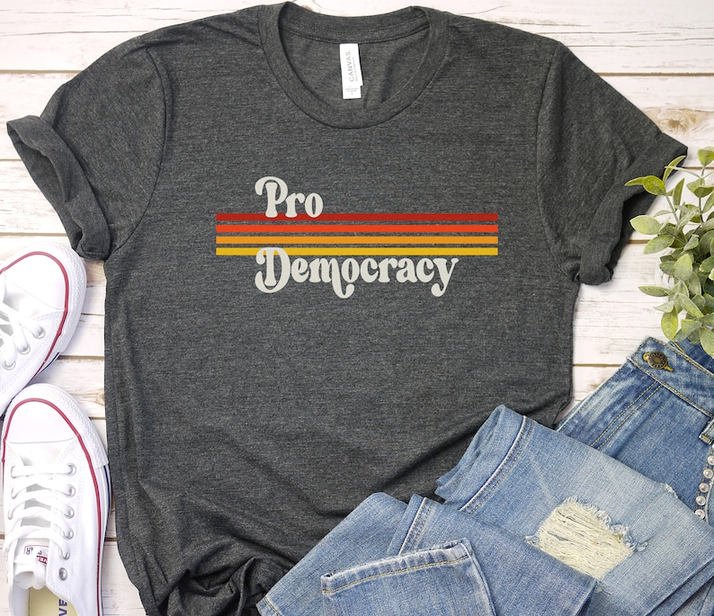 K&ouml;nnte beinhalten: Dunkelgraues T-Shirt mit dem Schriftzug "Pro Democracy" in Retro-Schrift, &uuml;ber drei horizontalen Streifen in Rot, Orange und Gelb. Das Shirt wird mit wei&szlig;en Turnschuhen und blauen Jeans gezeigt.