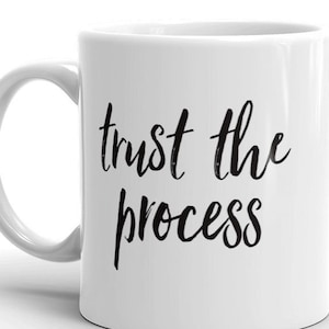 Op de afbeelding: Witte keramische koffiemok met zwarte tekst die "trust the process" luidt.
