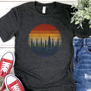 Pu&ograve; includere: T-shirt grigio scuro con grafica circolare di un tramonto su una foresta. Il design del tramonto include fasce di rosso, arancione, giallo e blu. La silhouette della foresta &egrave; blu scuro. La maglietta &egrave; abbinata a scarpe da ginnastica rosse e jeans blu.