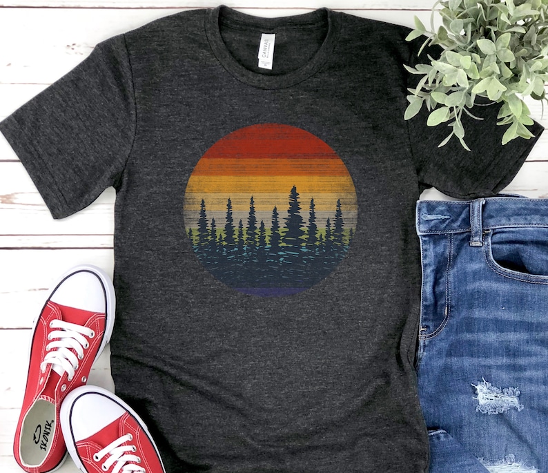 Pu&ograve; includere: T-shirt grigio scuro con grafica circolare di un tramonto su una foresta. Il design del tramonto include fasce di rosso, arancione, giallo e blu. La silhouette della foresta &egrave; blu scuro. La maglietta &egrave; abbinata a scarpe da ginnastica rosse e jeans blu.